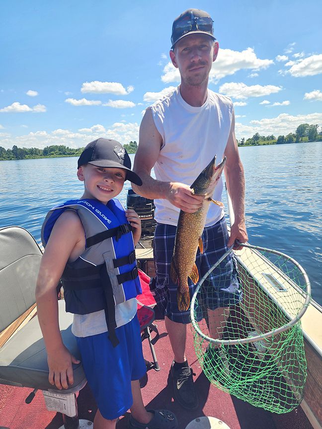 Two people on a boat hold a fish and a landing net on a sunny lake.