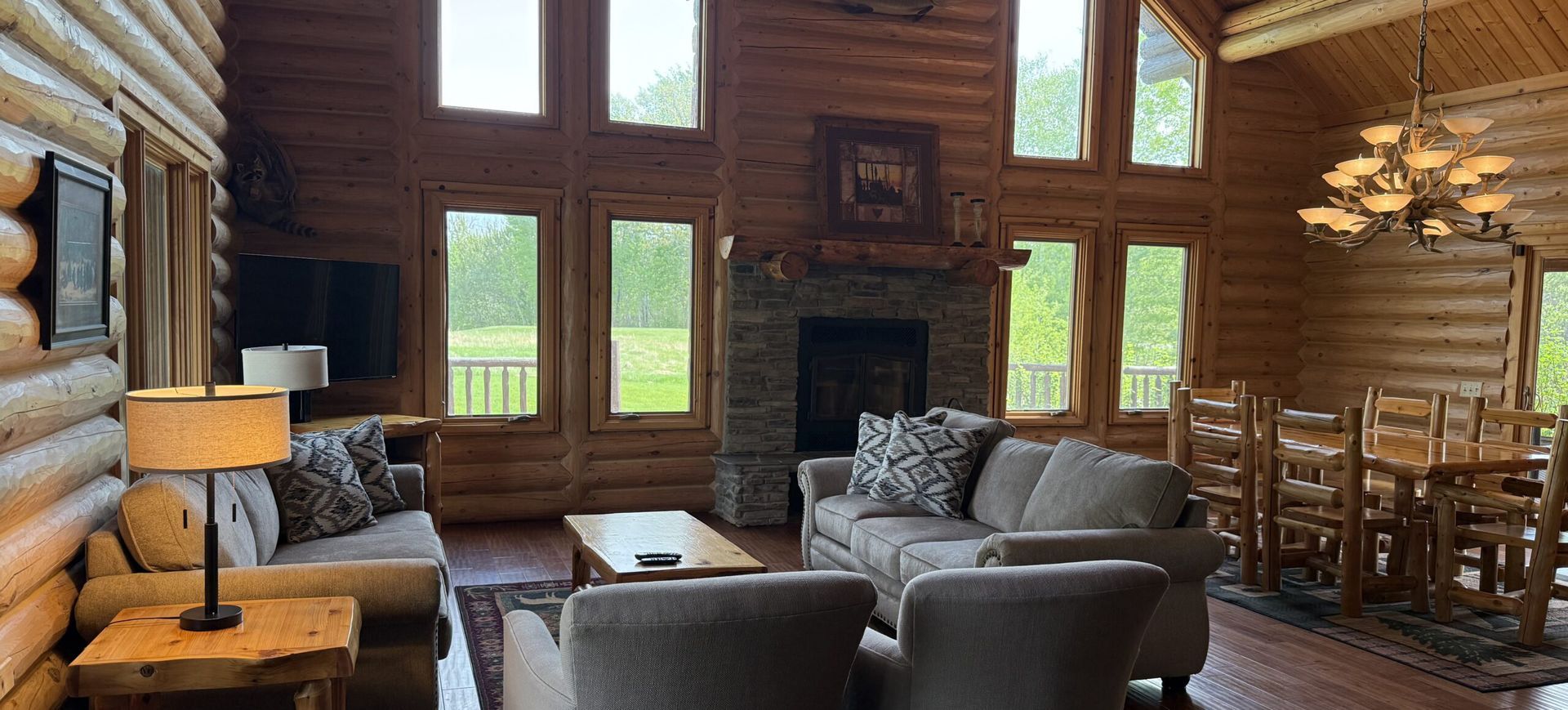 Cozy log cabin living room with large windows, fireplace, gray chairs, and wooden dining table