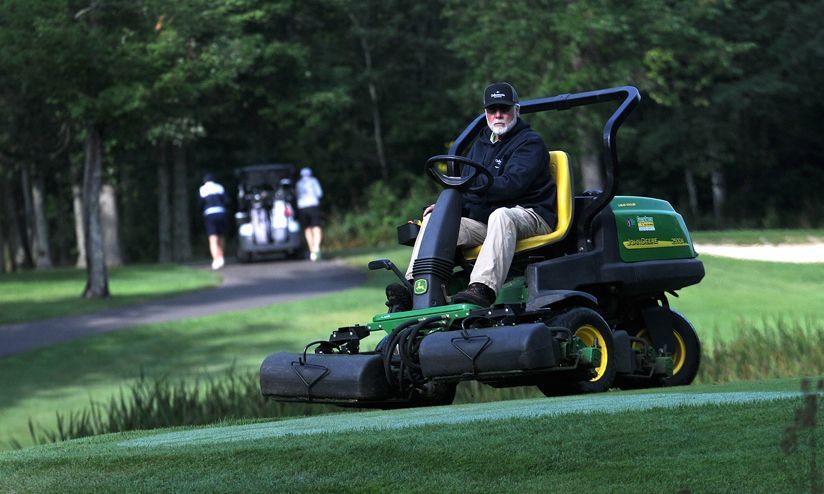 Person driving a green riding mower on a grassy golf course with trees in the background