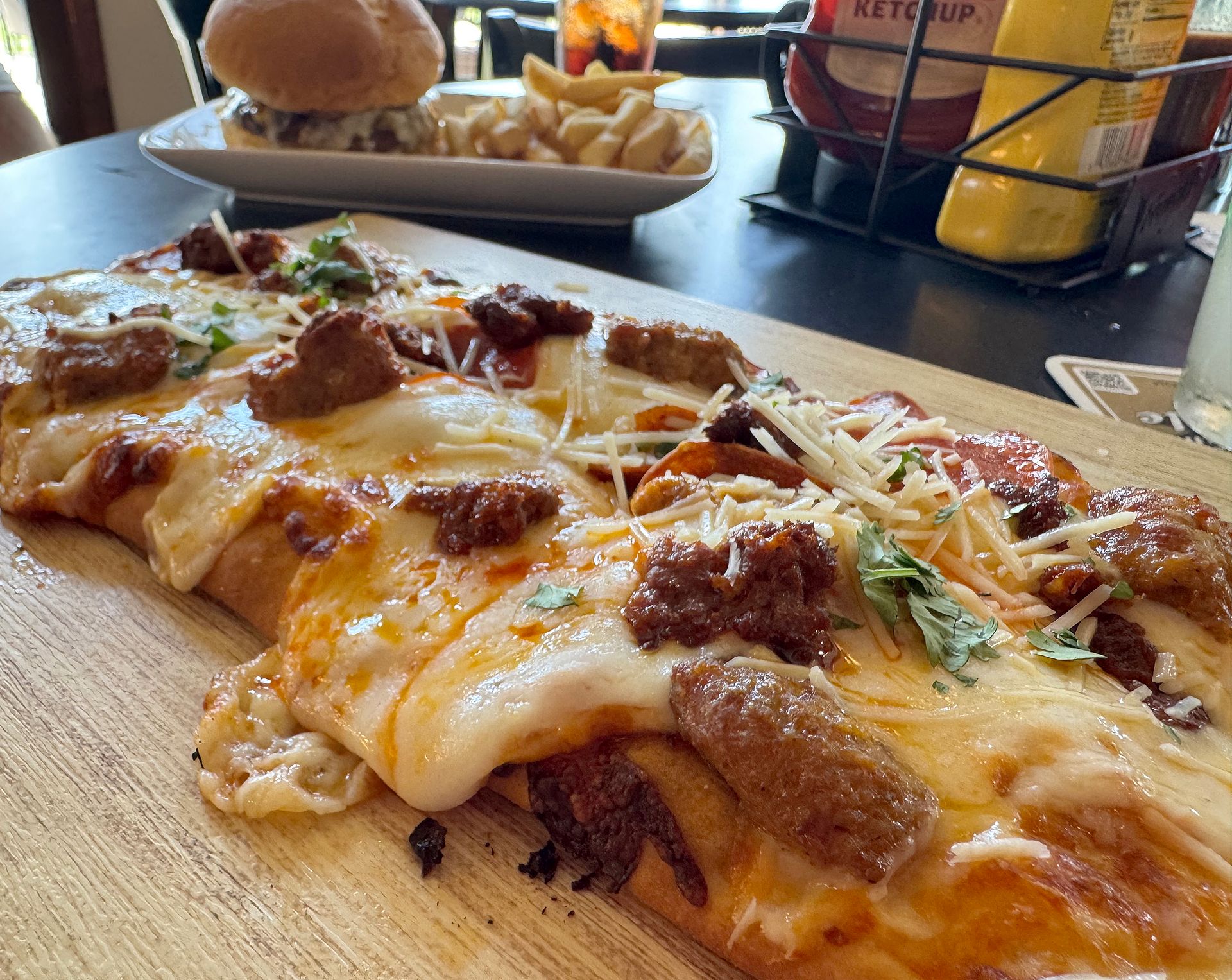 Cheesy meat pizza slices on a wooden board beside a burger basket in a restaurant