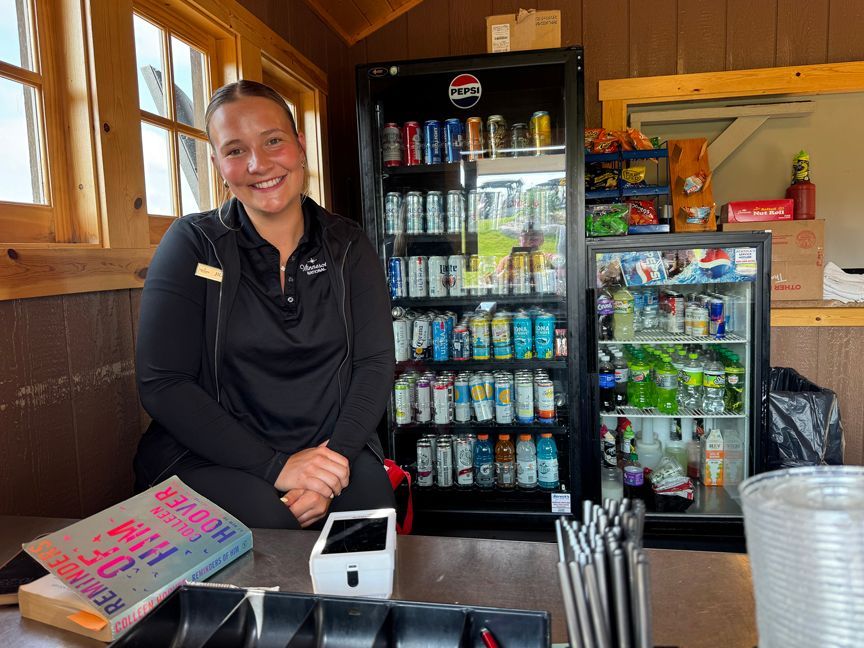 Smiling vendor behind a counter with a drink fridge in a small wooden kiosk