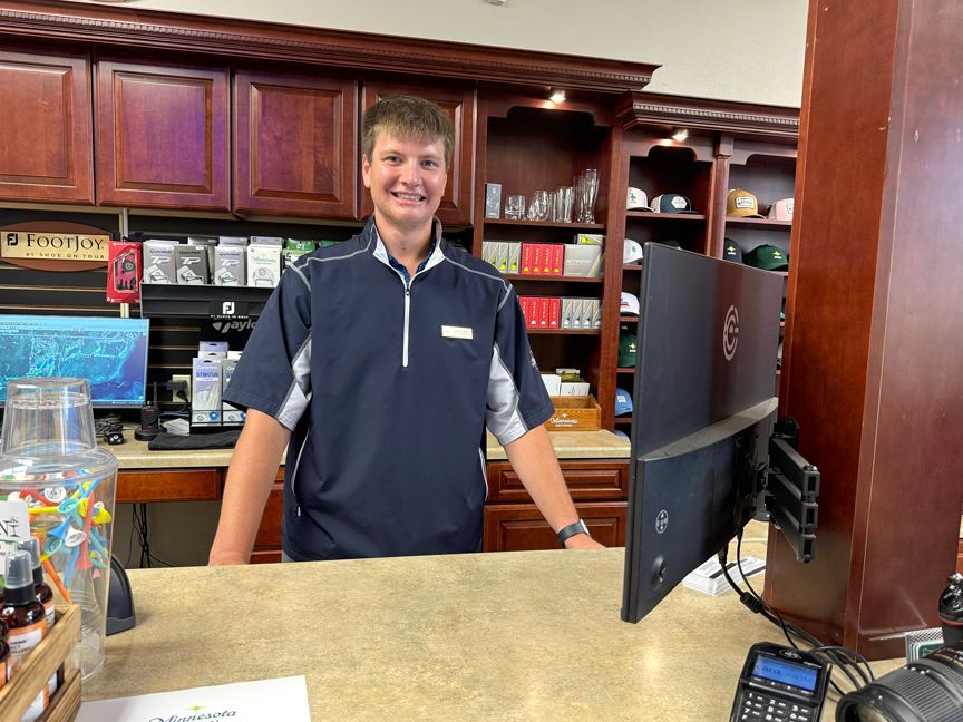 Smiling employee behind a café counter, with wood cabinets and a computer monitor nearby.