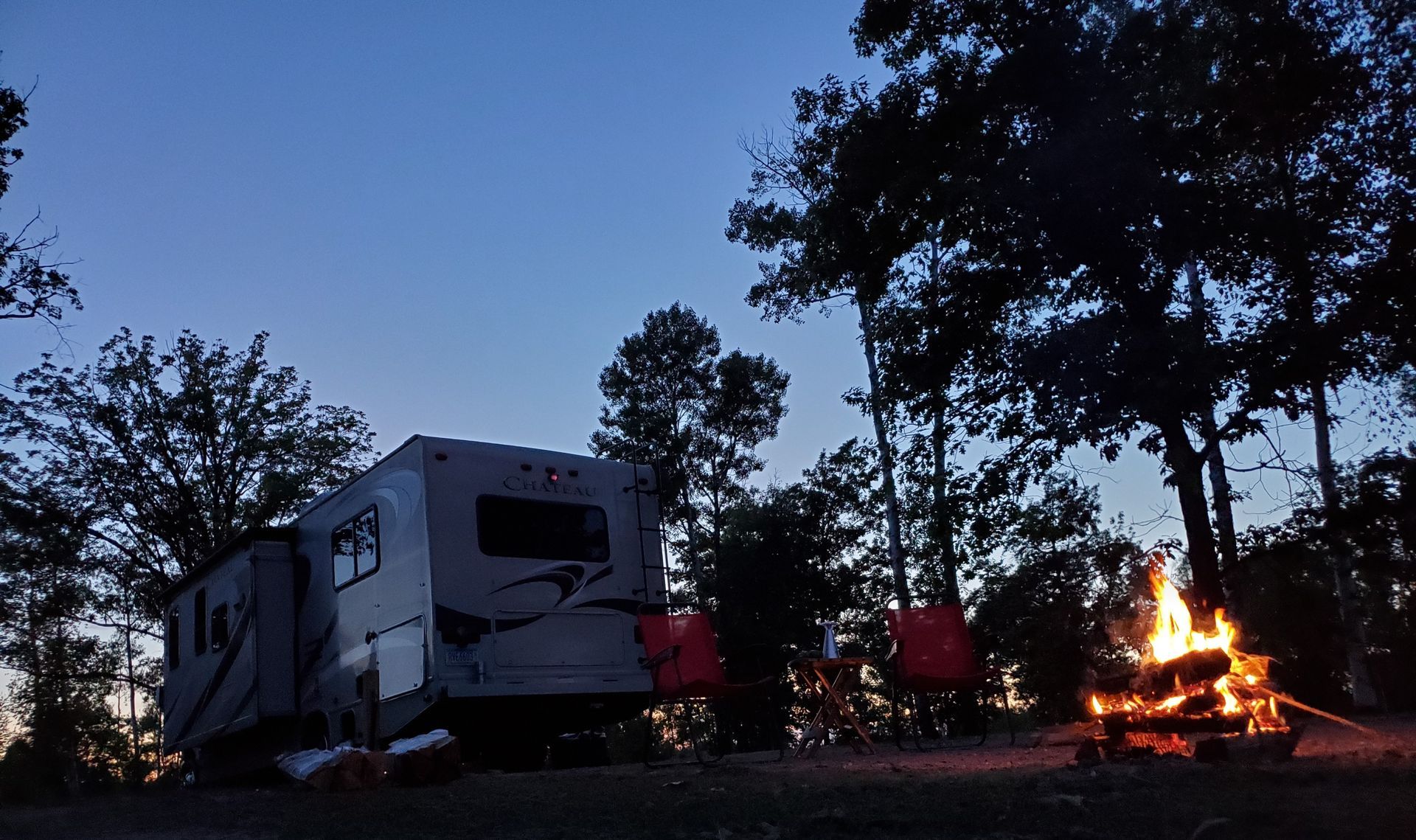 Campsite at dusk with an RV, trees, and a small campfire glowing on the right.