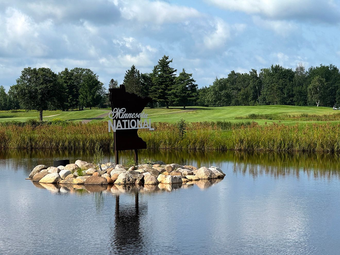 A sign reading “Welcome Matic” on a rock island in a calm pond, with grassy fields and trees behind.