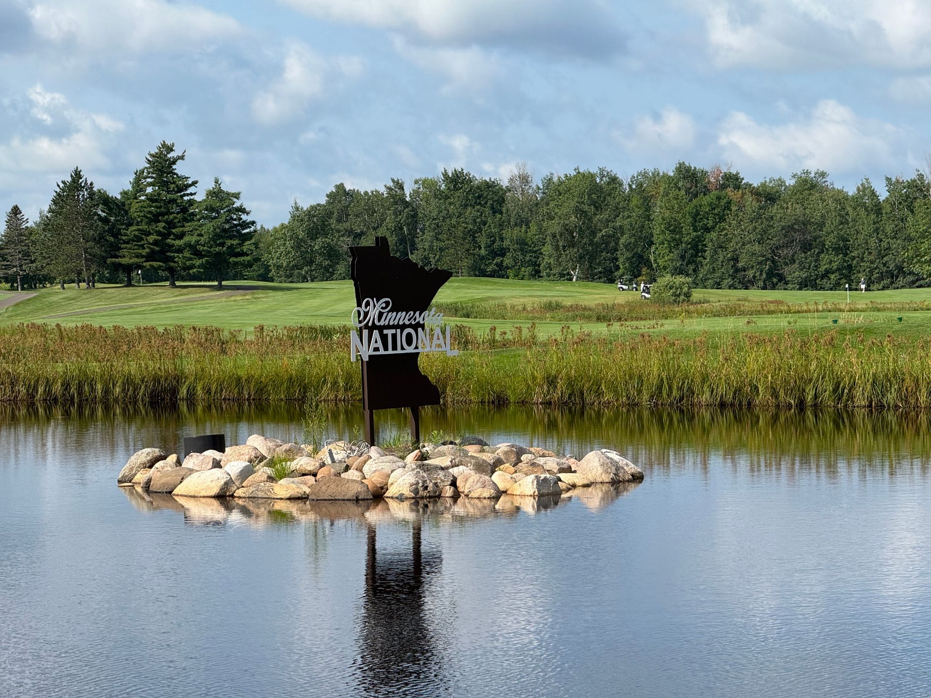 Small rock island in a pond with a black national park sign, surrounded by green grass and trees.