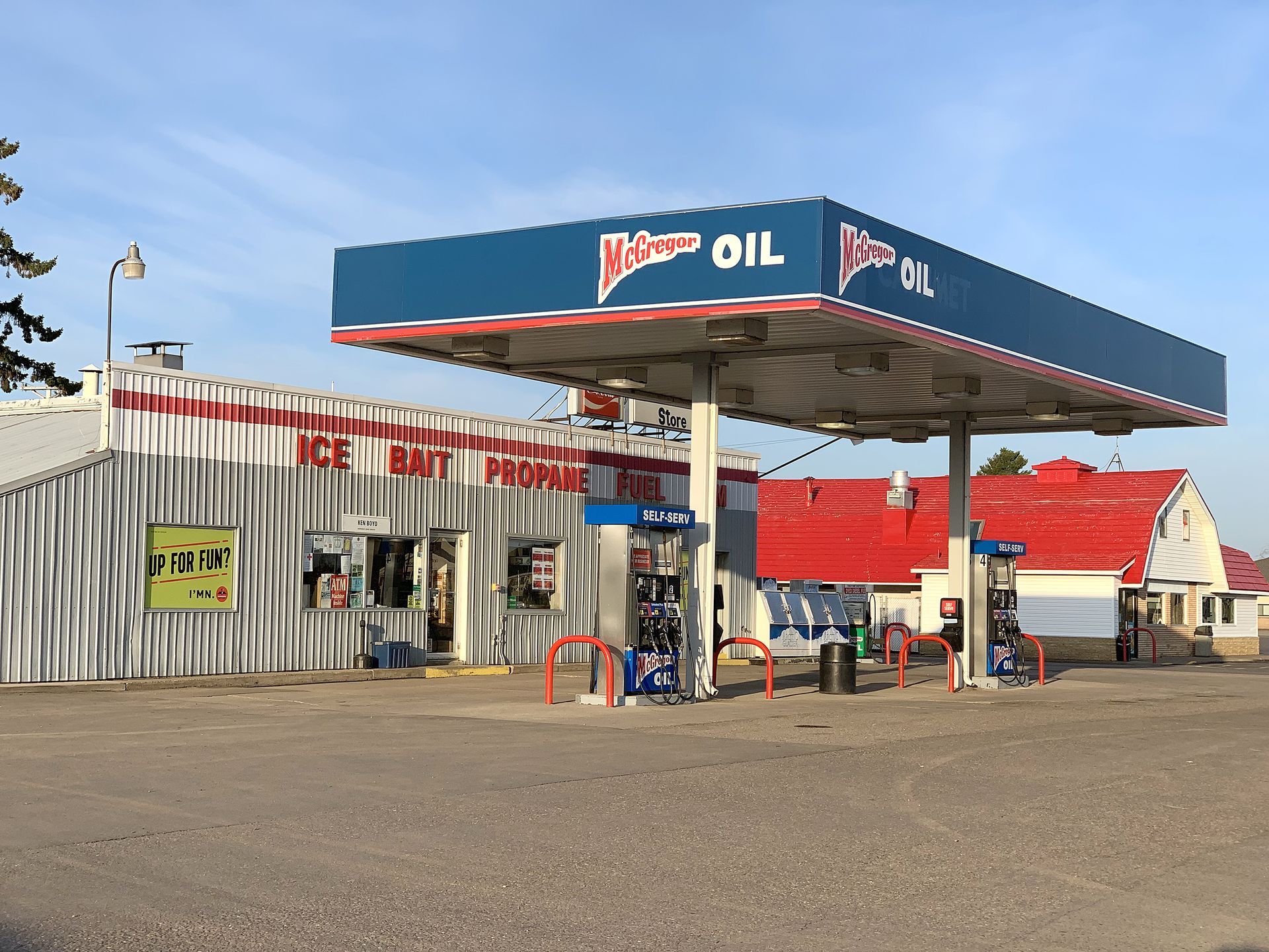 Small roadside gas station with a blue canopy and red-roofed buildings under a clear sky.