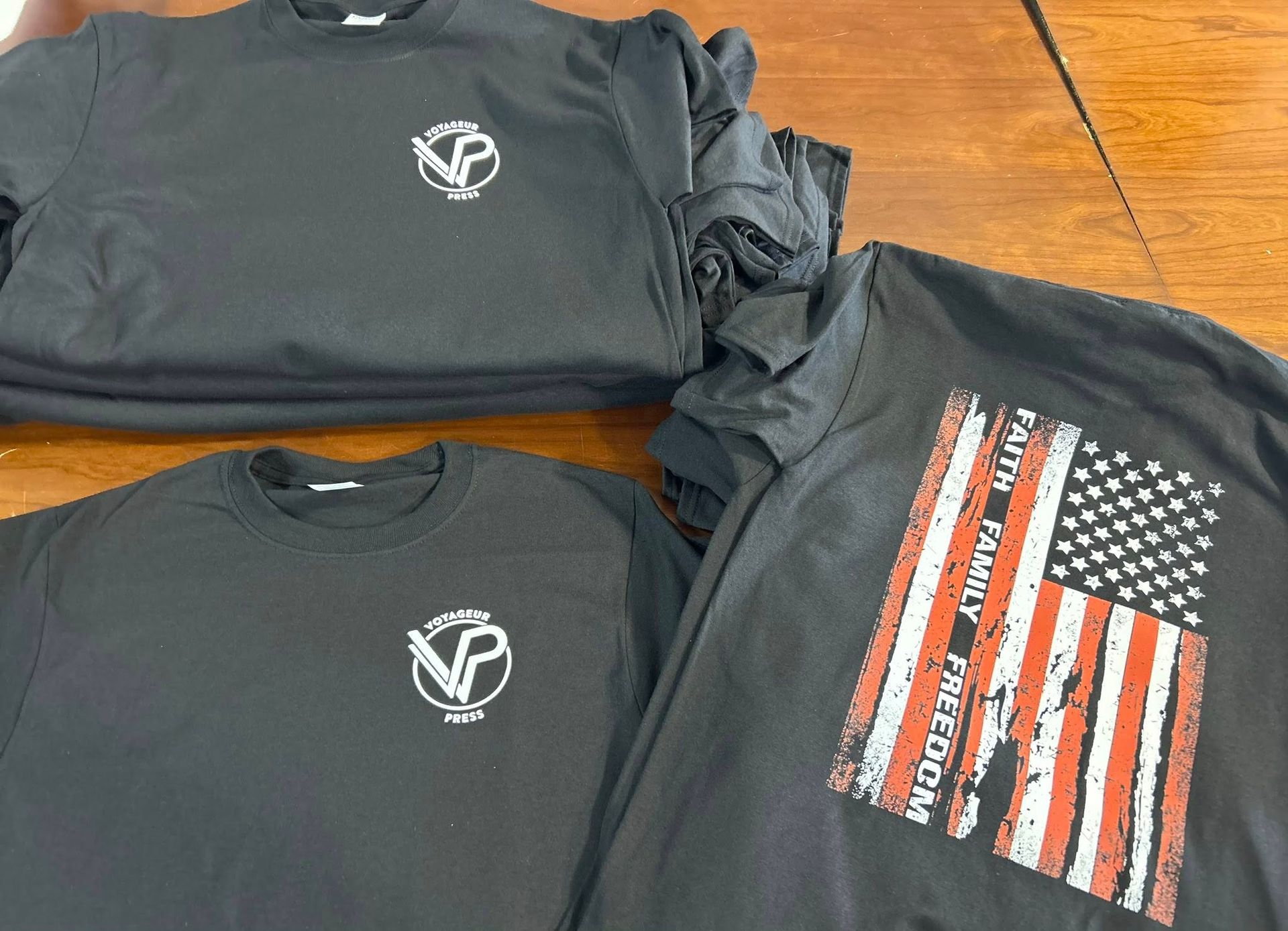Three black shirts and a gray top with small logos and one American flag graphic, laid on a wooden table