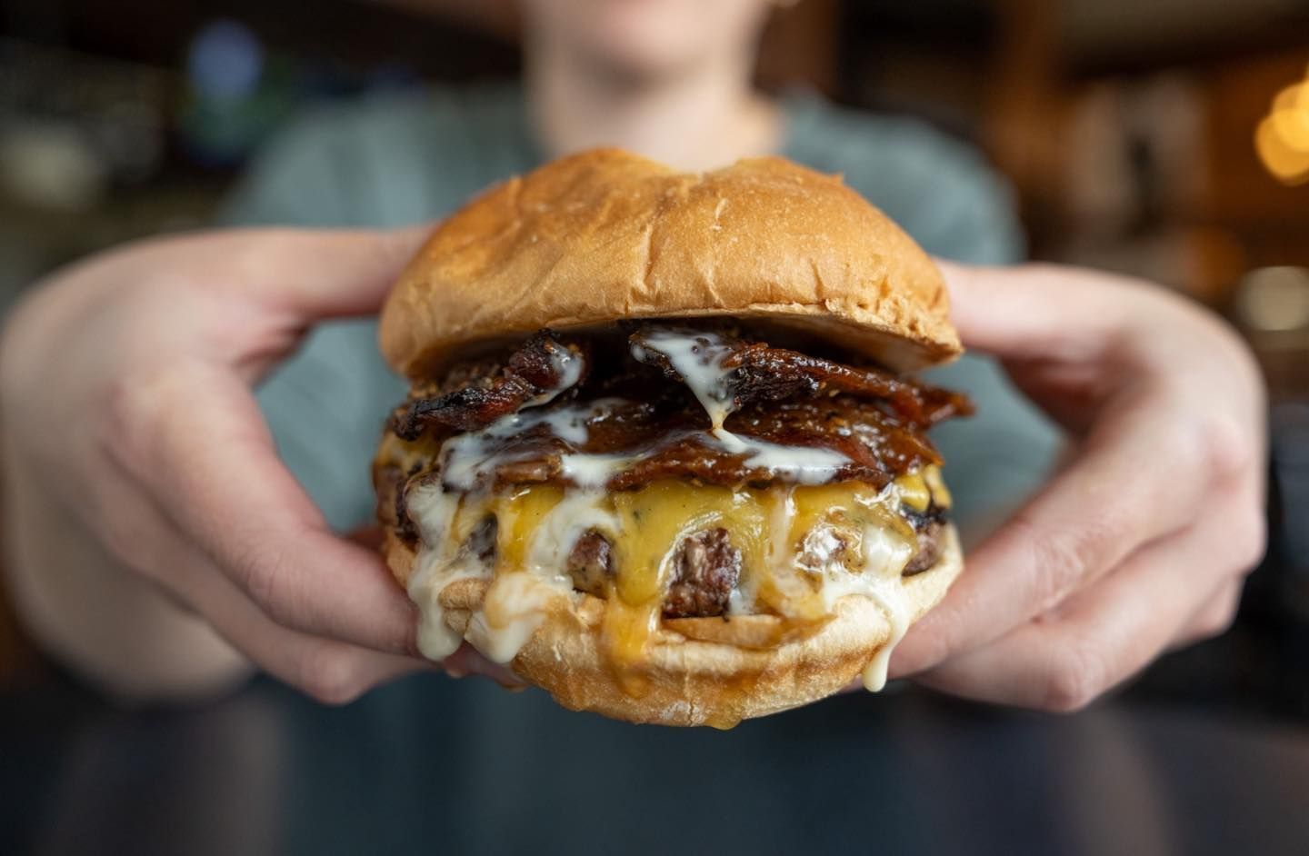 Hands holding a bacon cheeseburger with melted cheese and dark sauce on a bun