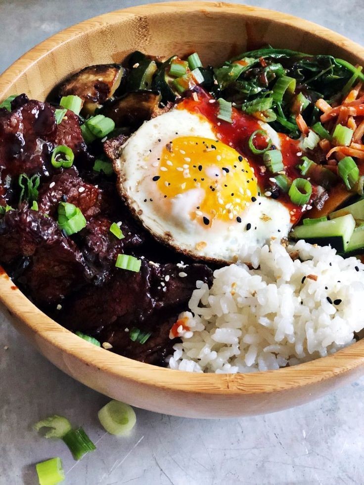 Rice bowl with grilled beef, fried egg, greens, and chopped green onions in a wooden bowl