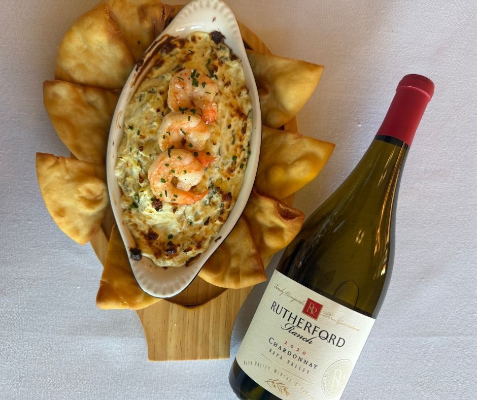 Baked seafood dip topped with shrimp, surrounded by fried bread, beside a bottle of white wine