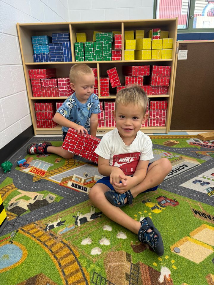 Two children sitting on a classroom rug near cubbies, playing with toys and books.