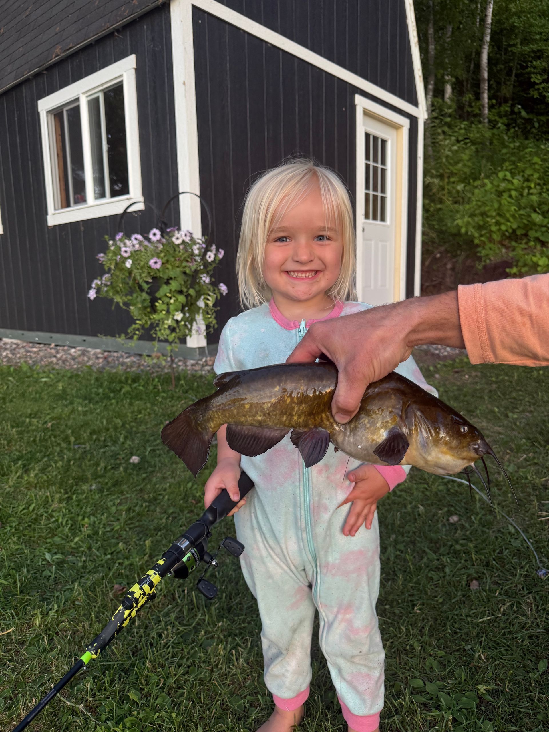 Child smiling beside a house while holding a fish with an adult’s hand supporting it