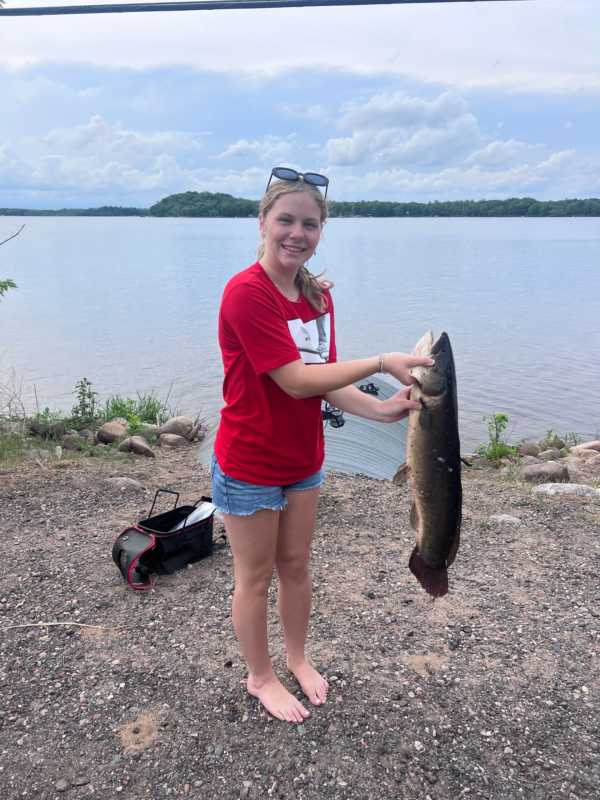 Person holding a large fish by a lakeshore under a cloudy sky