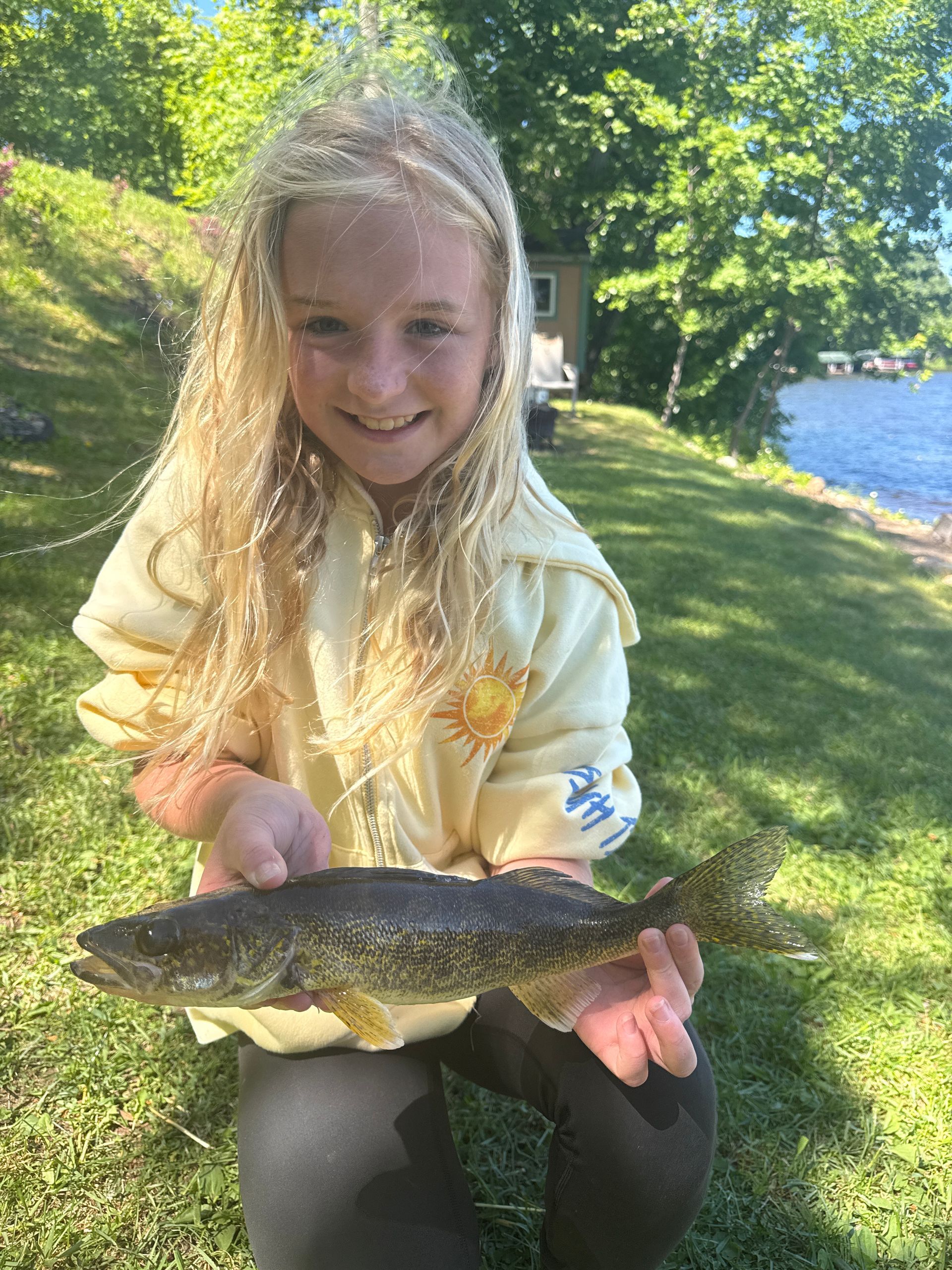 Smiling person holding a large fish outdoors by a lake, with trees and bright sunlight