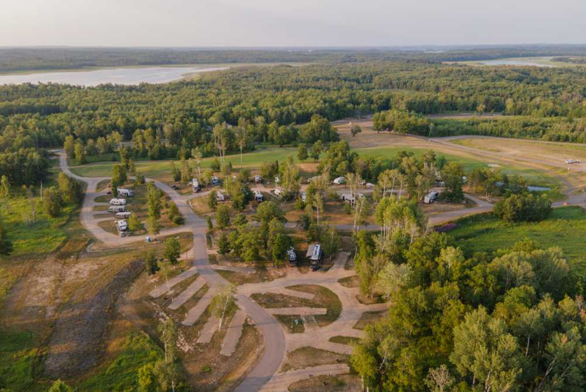 Aerial view of a wooded campground with winding roads, scattered RVs, and a large open field.