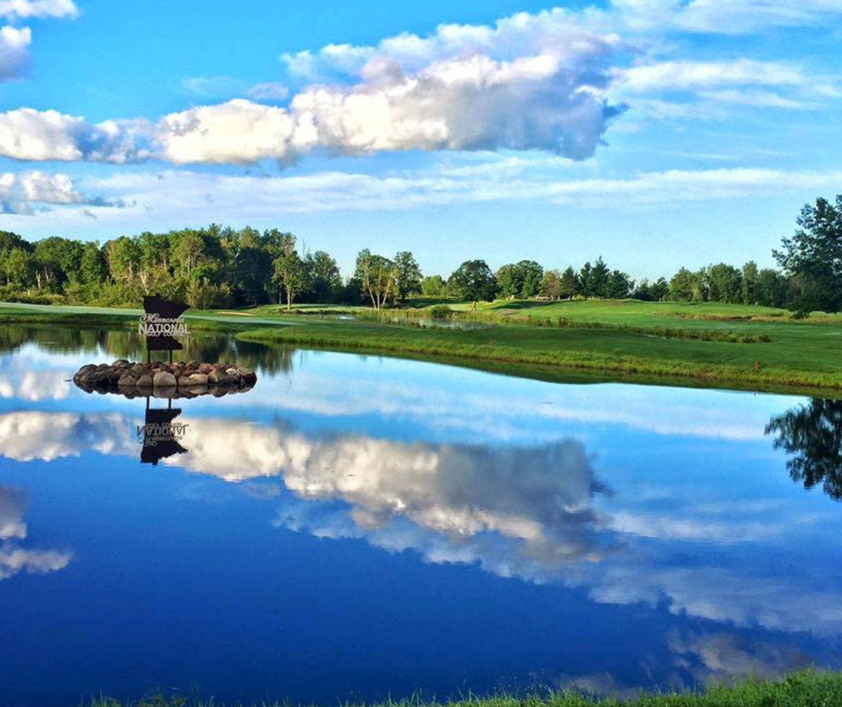 Calm lake reflecting blue sky, clouds, and green trees beside a small fountain and grassy shore