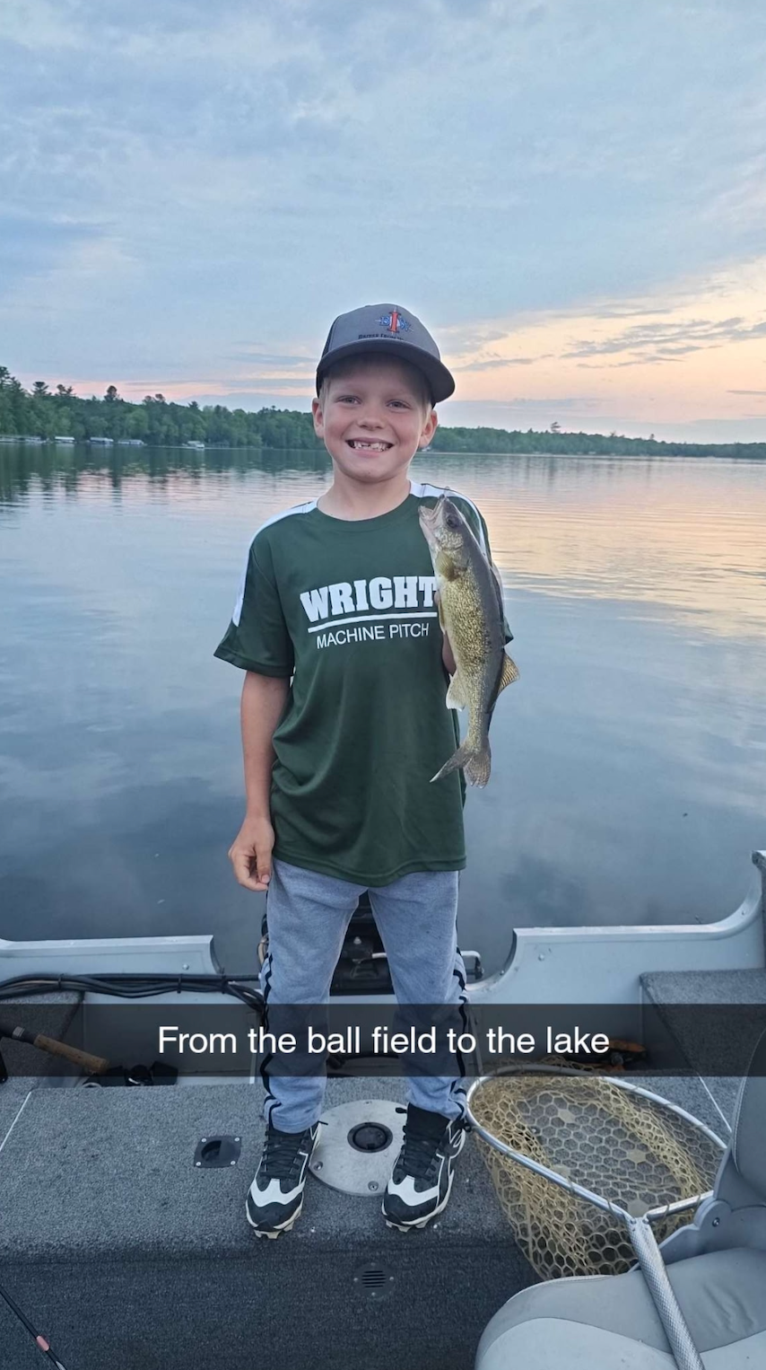 Boy smiling on a boat holding a fish, with a calm lake and sunset in the background.