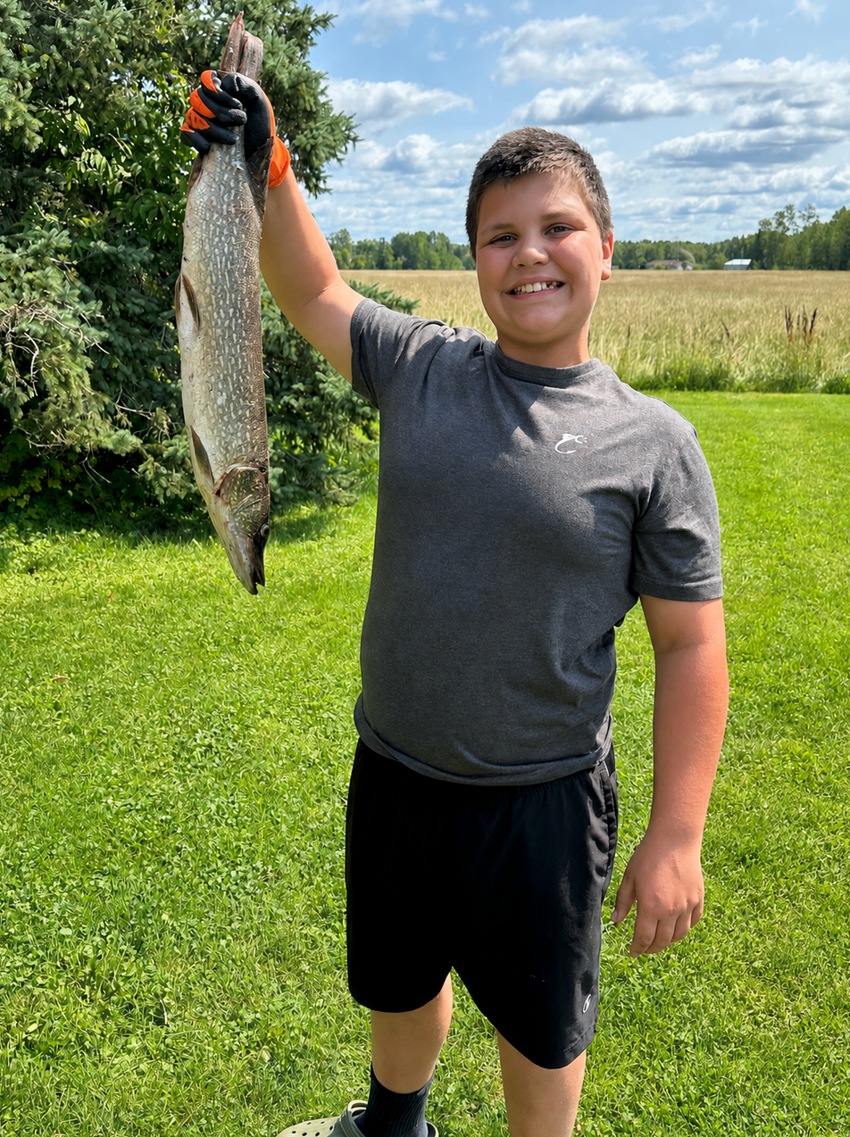 Person standing on grass, smiling and holding up a large fish by a lake under a cloudy sky