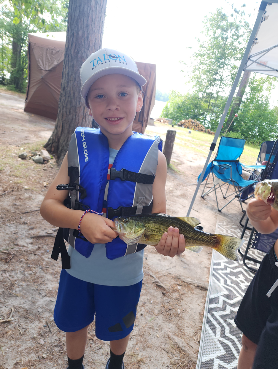 Child in a blue life jacket and cap holding a fish outdoors by a campsite.