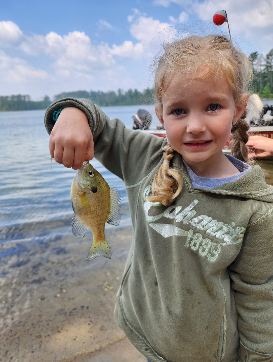 Smiling child holding up a small fish by a lakeshore, with water and sandy beach in the background