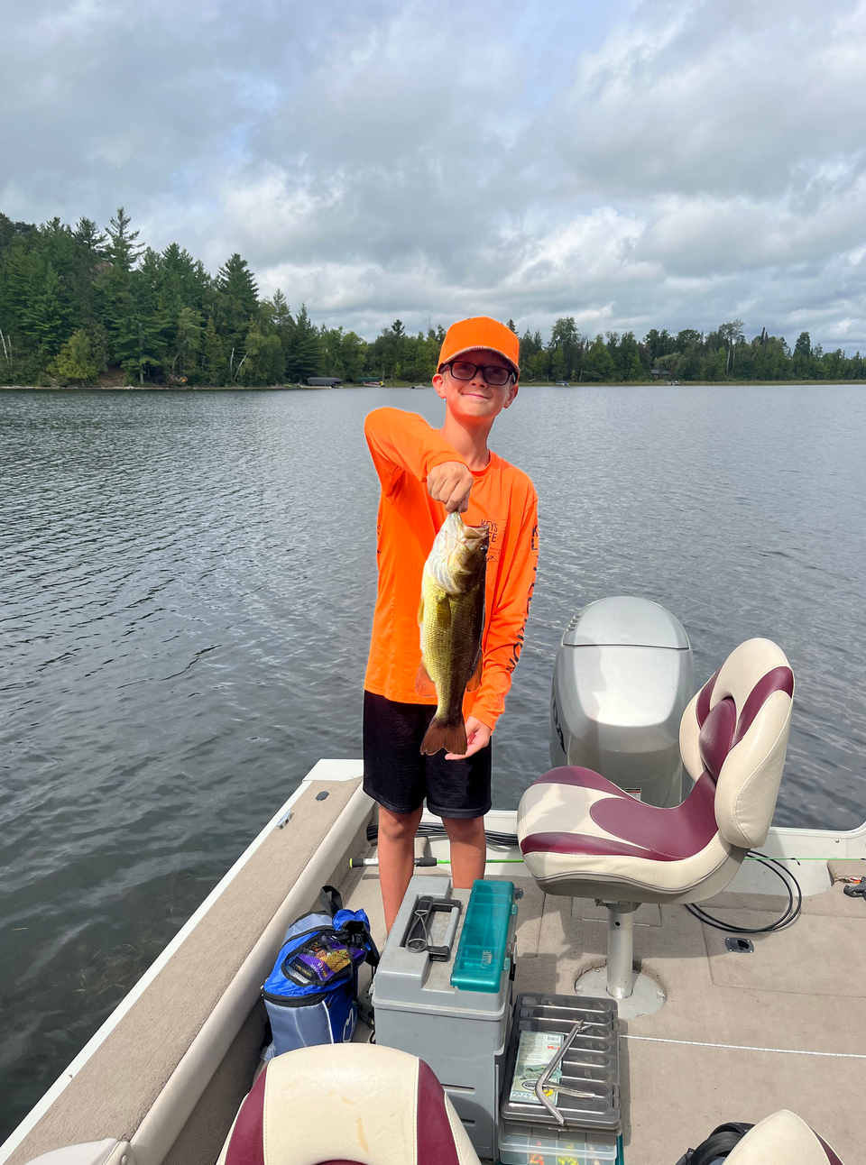 Person in orange shirt and cap holds up a fish on a boat on a lake under cloudy skies.
