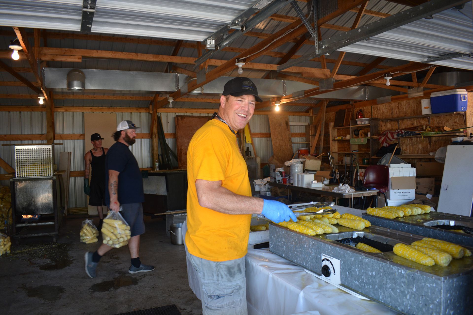 Workers preparing food on trays in a warehouse kitchen, with one man in a yellow shirt in the foreground.