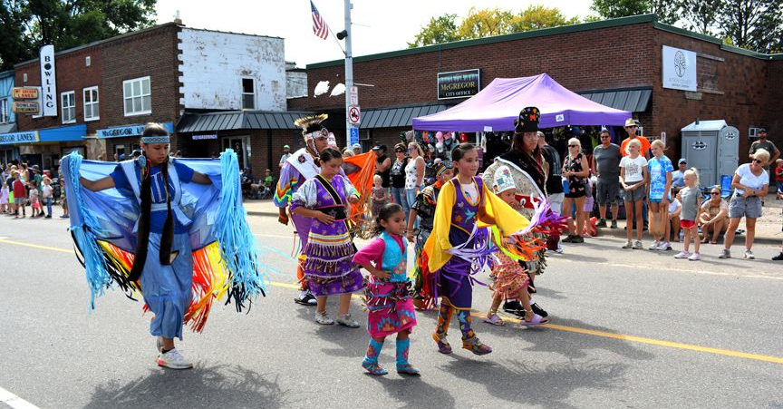 Dancers in colorful costumes perform in a street parade with spectators and buildings in the background