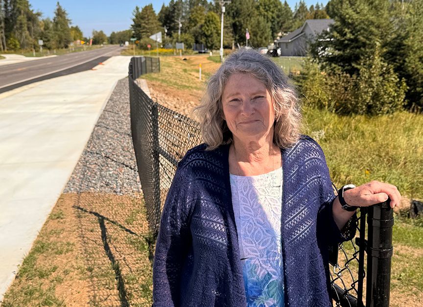 Woman standing by a fence on a sunny roadside, wearing a blue cardigan and white top.