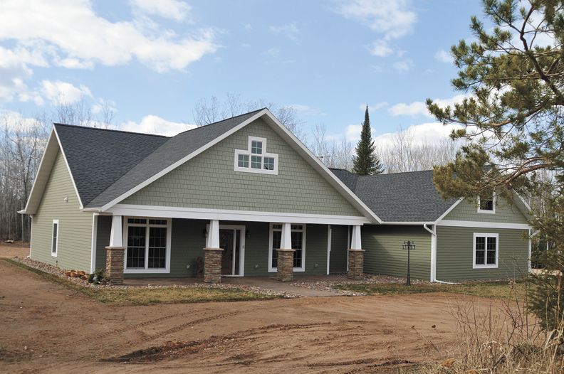 New gray house with green roof, white trim, and porch on a bare lot under a partly cloudy sky
