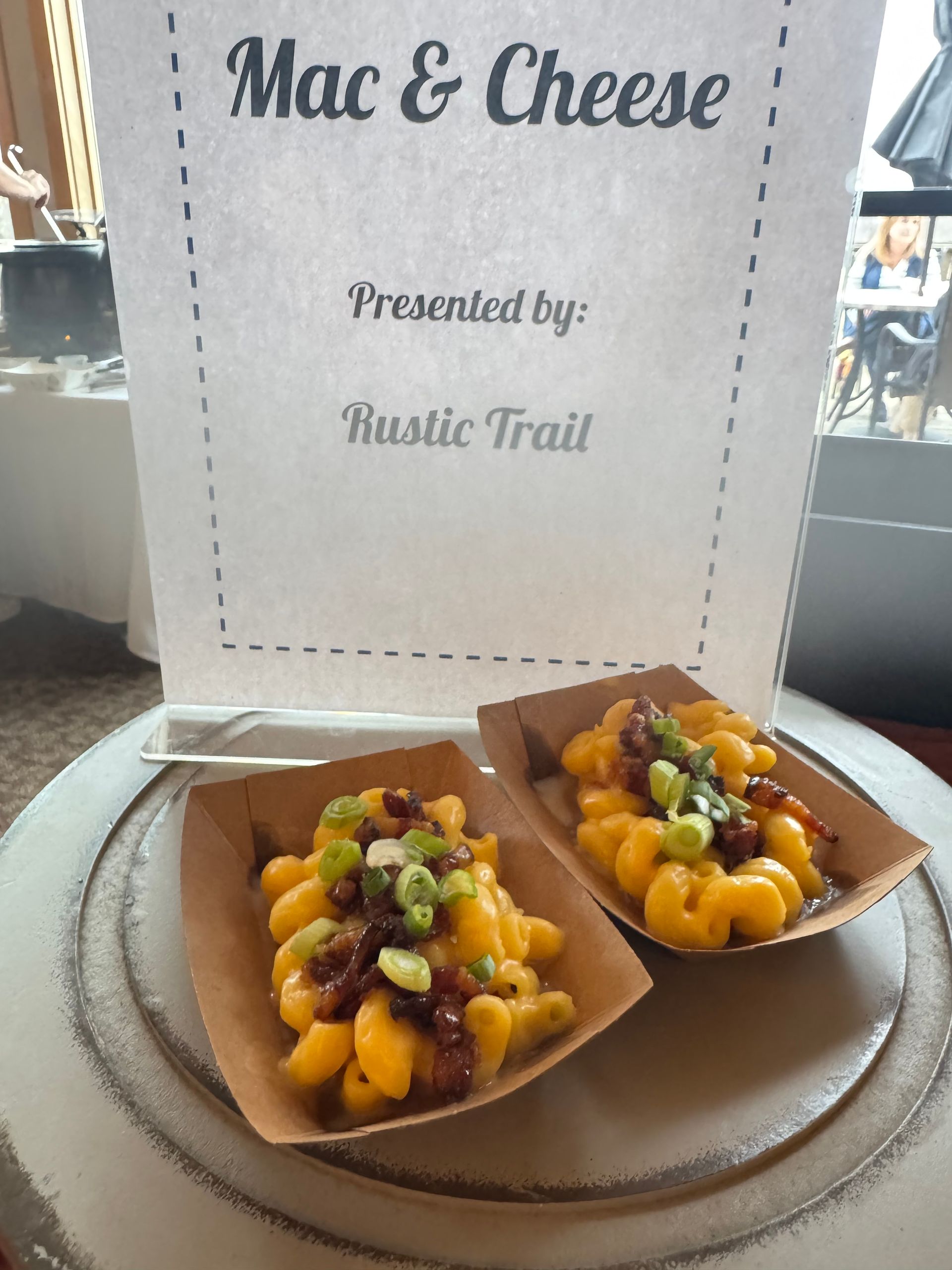 Mac and cheese samples in small cardboard trays on a plate, with a Rustic Trail sign behind.