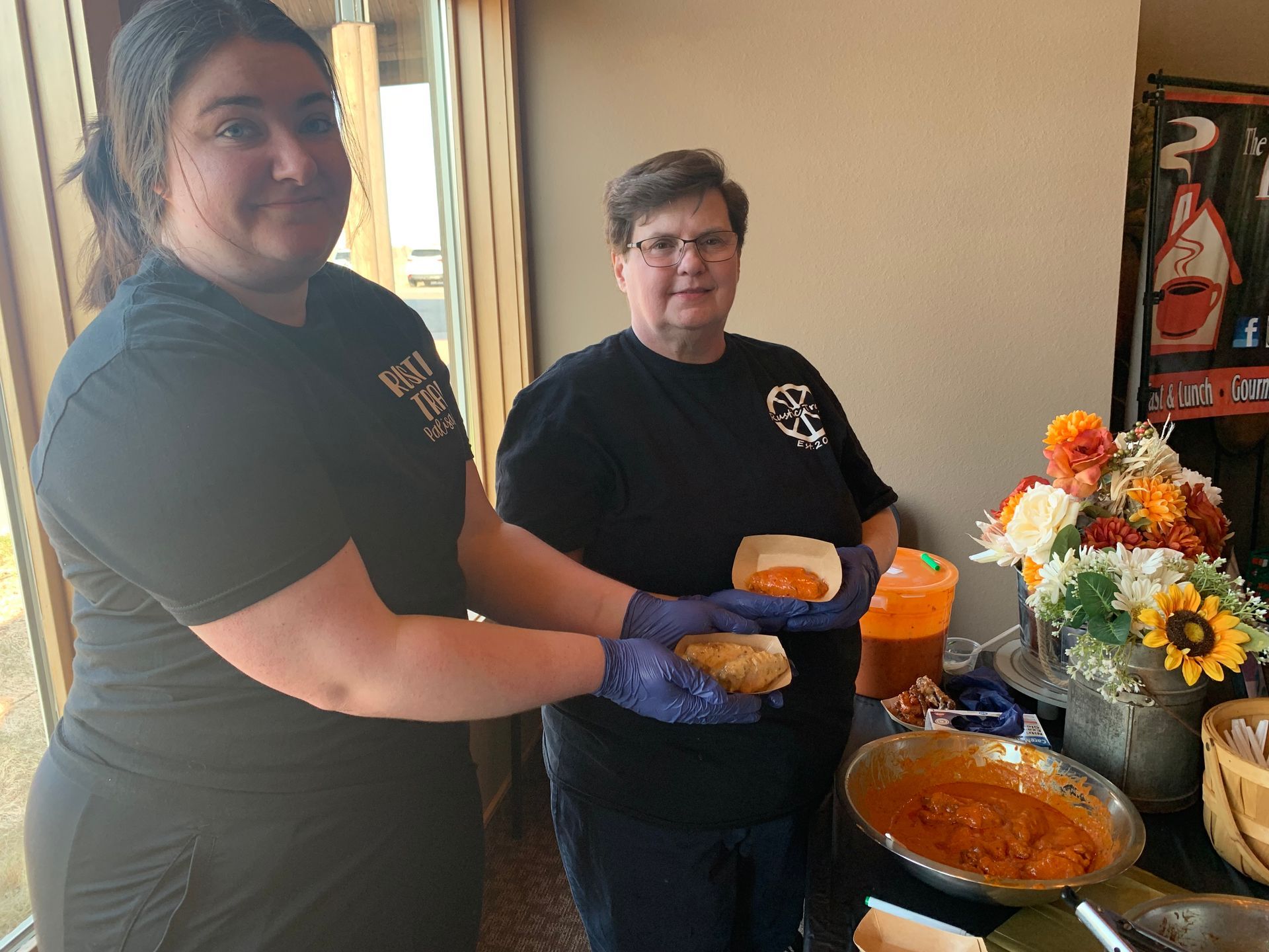 Two volunteers serve food at a buffet table with orange soup and flowers nearby.