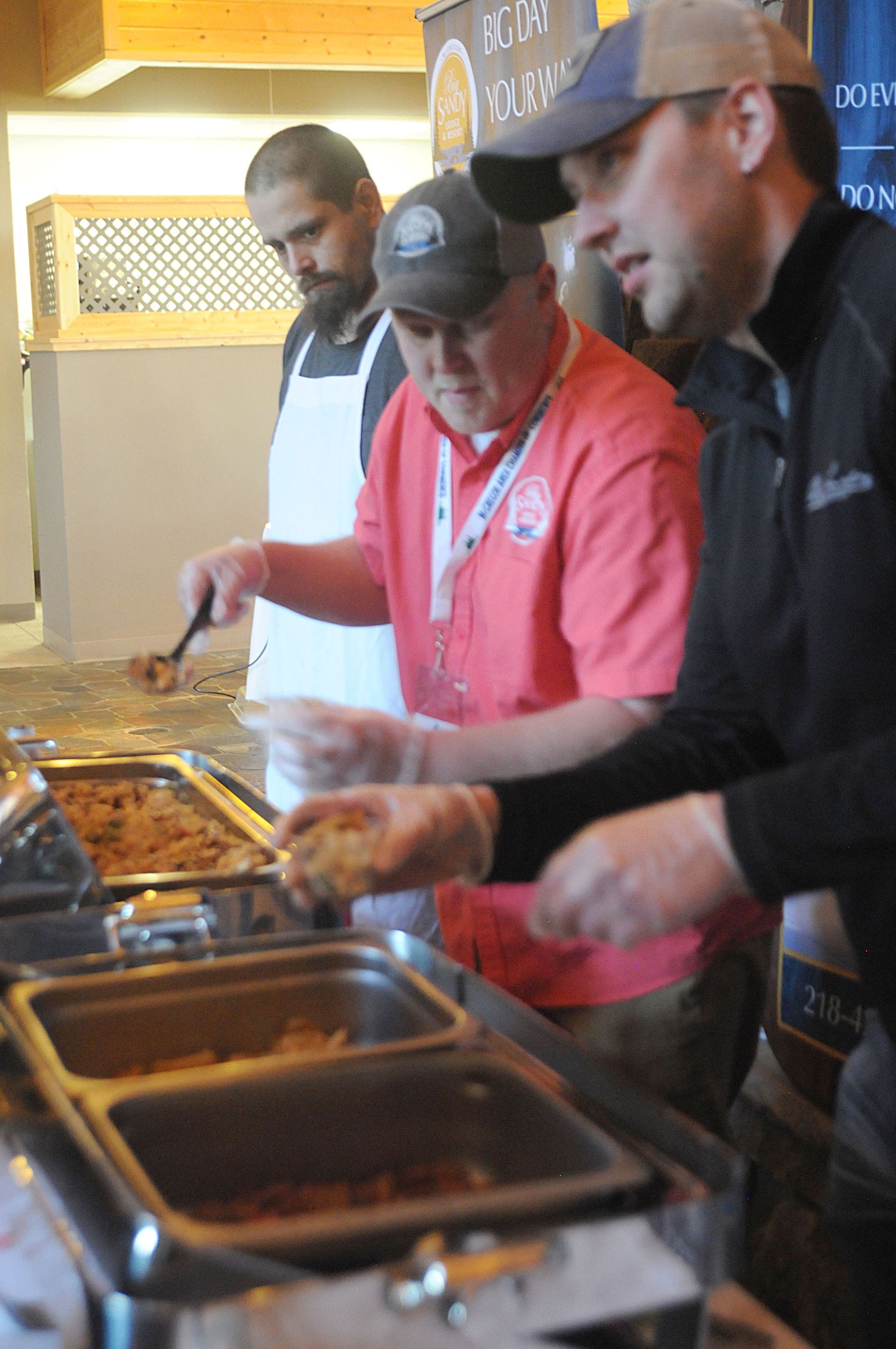 Three people serving food at a buffet counter with trays of hot dishes