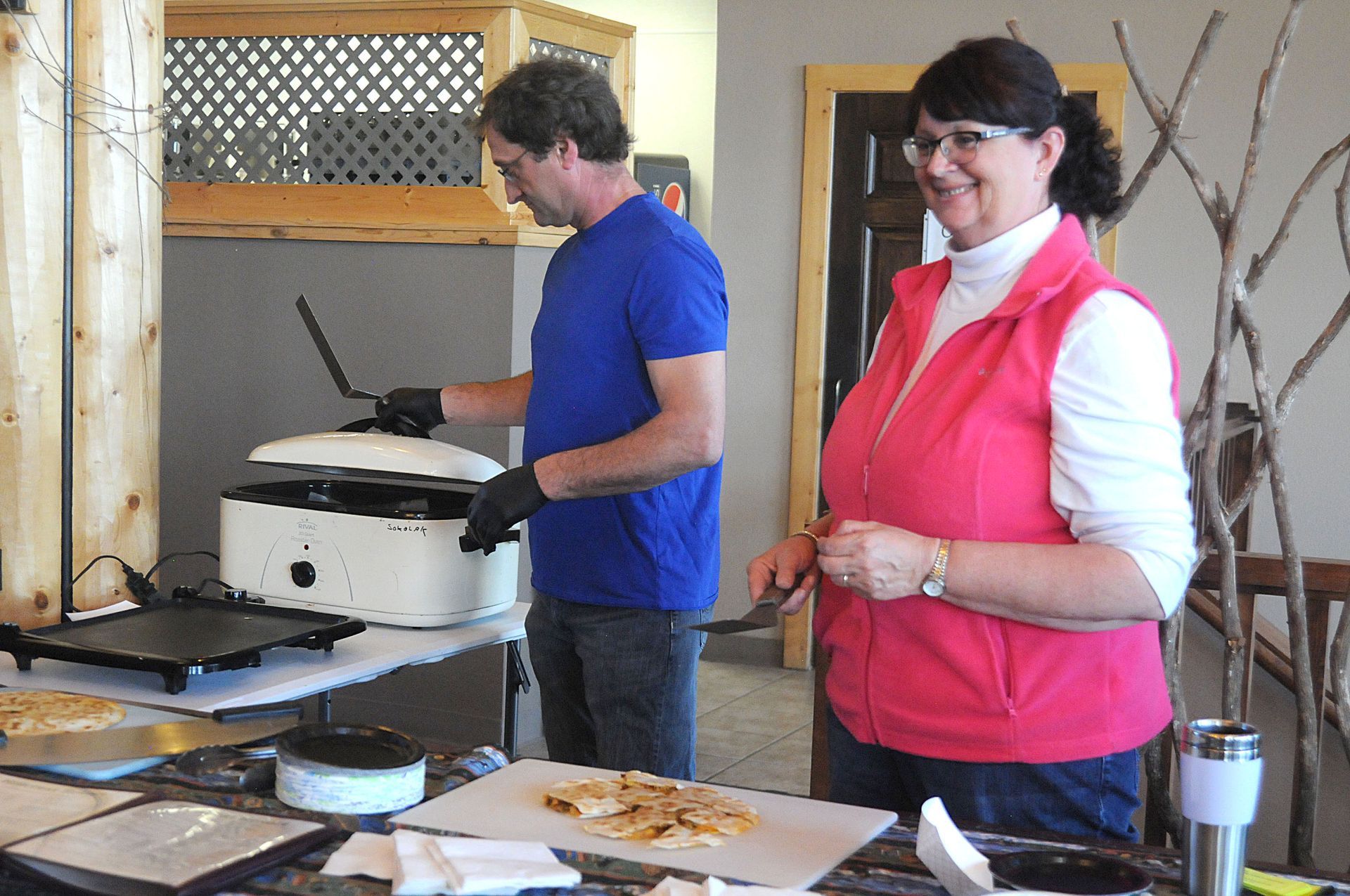 Two people at a buffet table, one cooking on a griddle and one smiling in a pink vest.