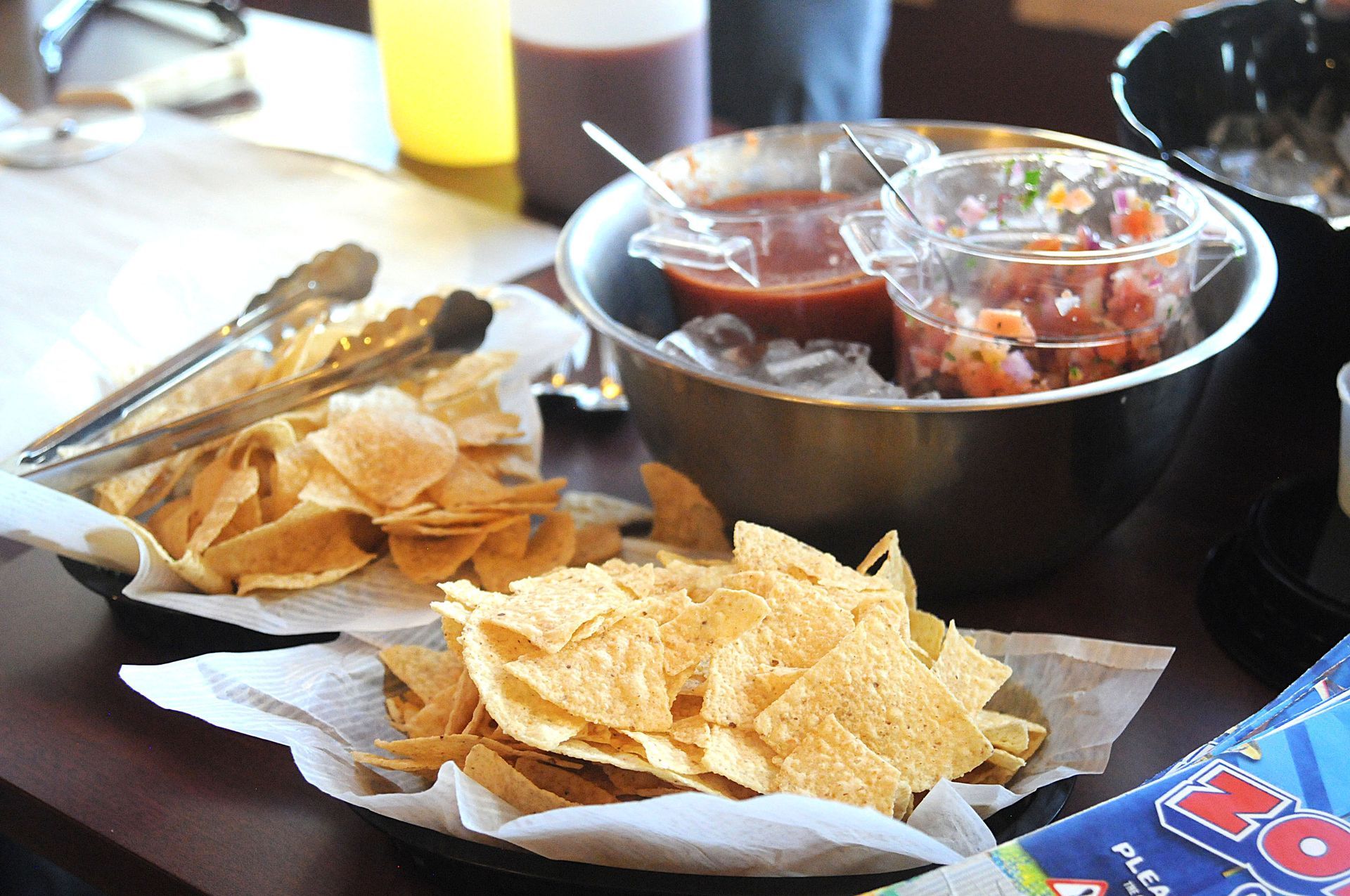 Tray with tortilla chips and dipping bowls on a table, with tongs and a drink in the background