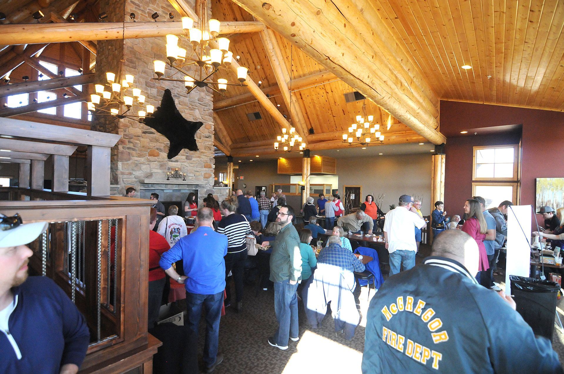 Crowded rustic lodge interior with people gathered under a wood-beam ceiling and chandelier.