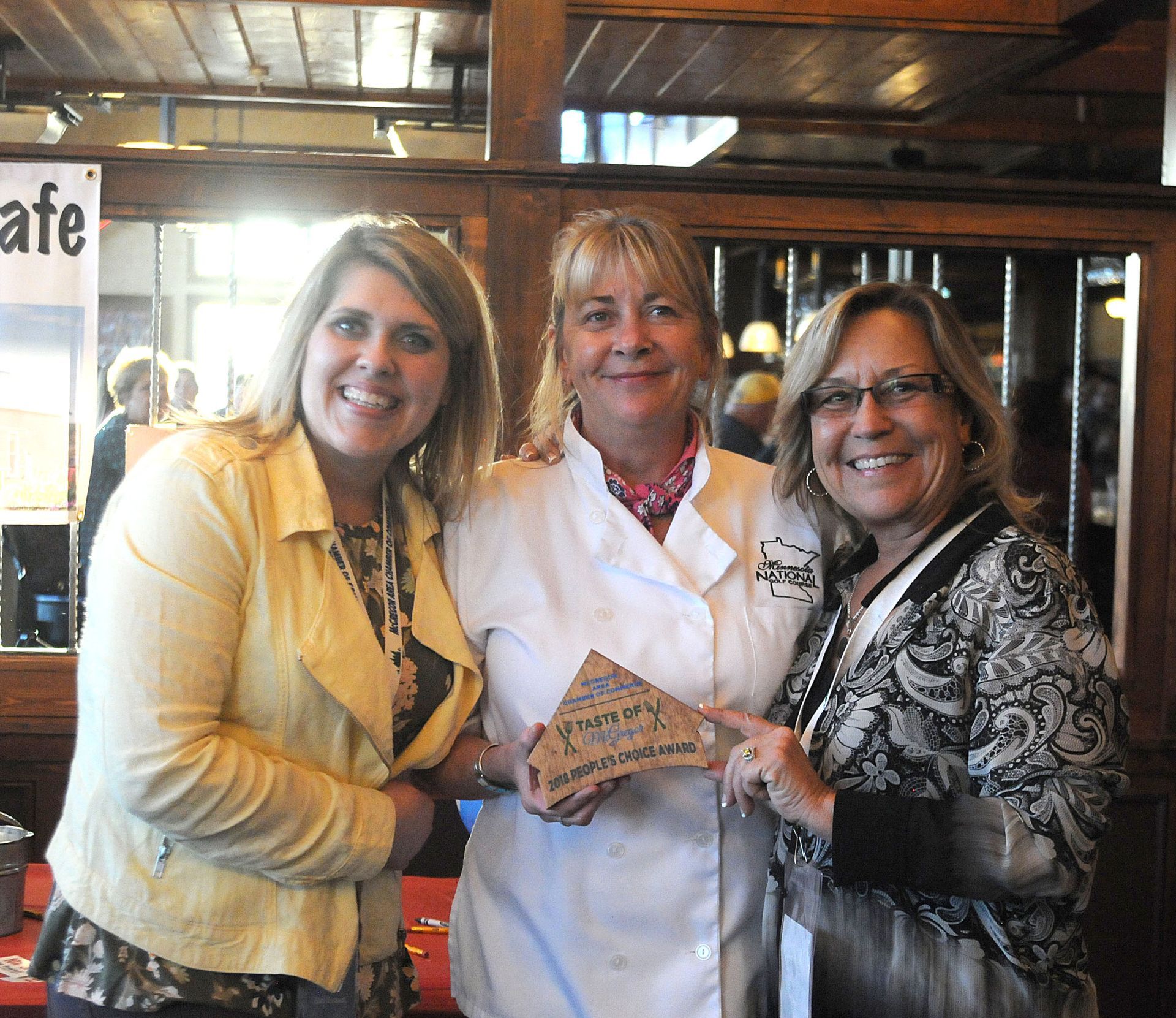Three women smiling indoors, holding a patterned object together in a rustic café.