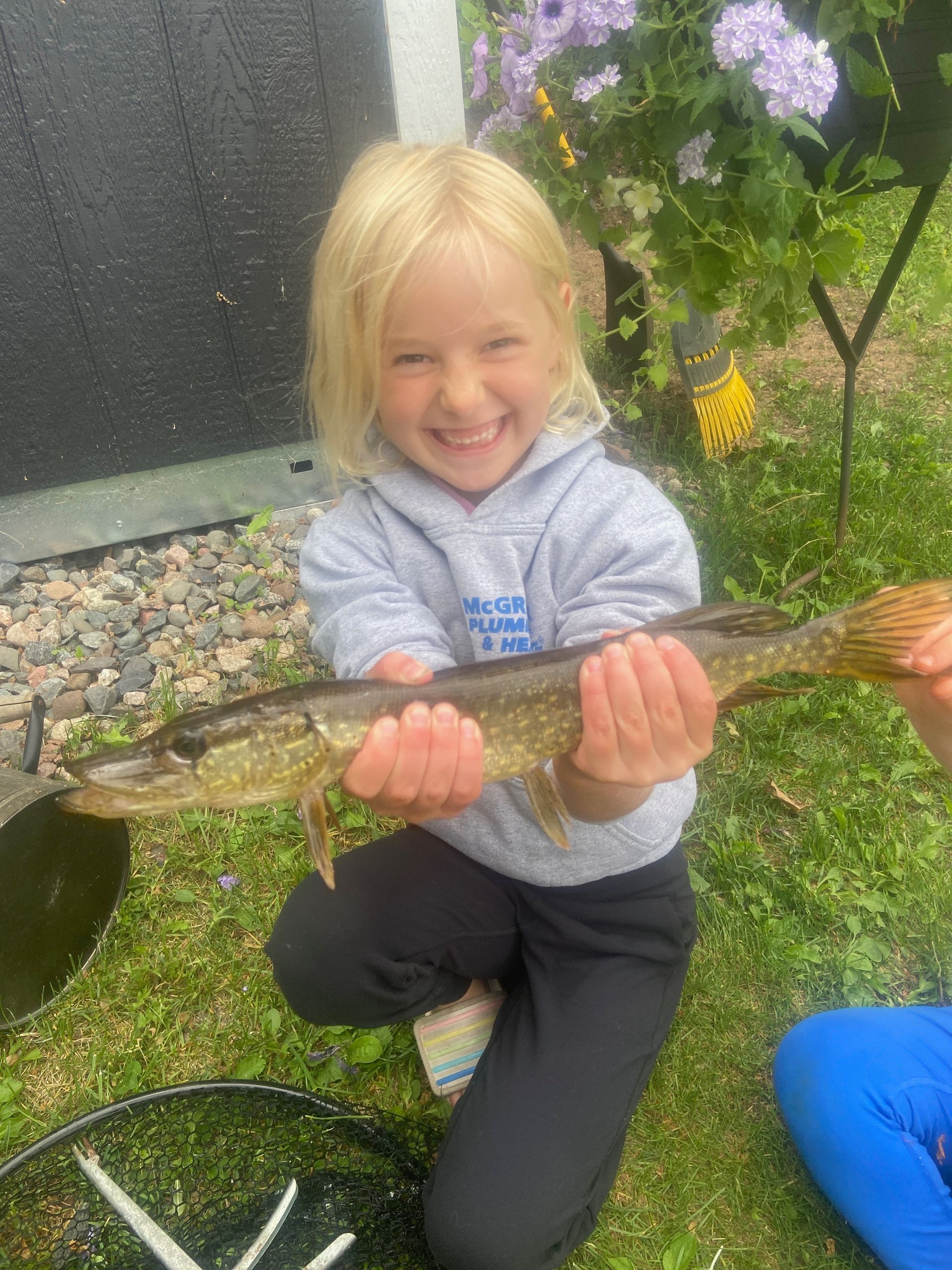 Child kneeling outdoors, smiling and holding a long fish in a garden.