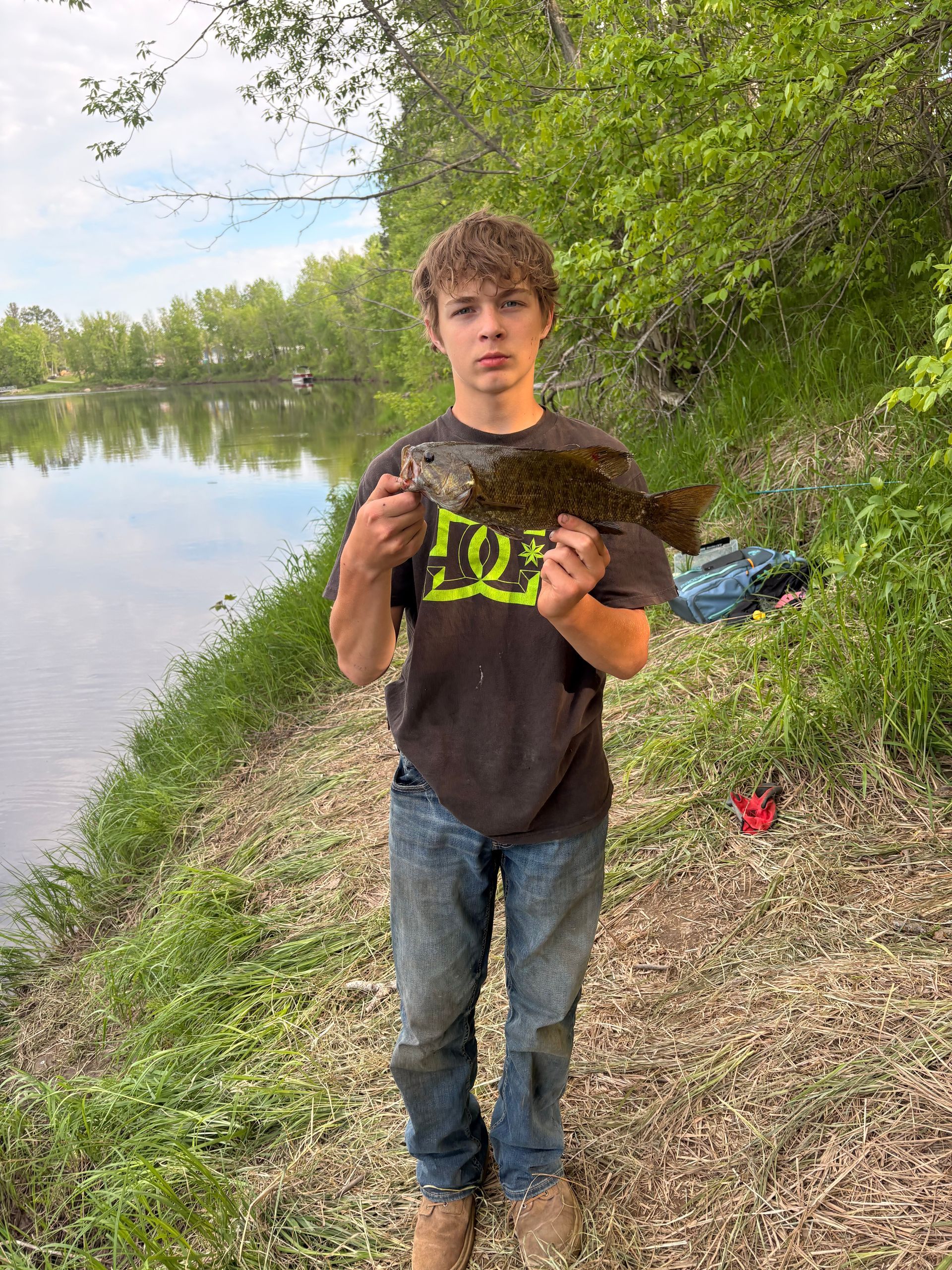 Person holding a large fish on a grassy riverbank near calm water and trees