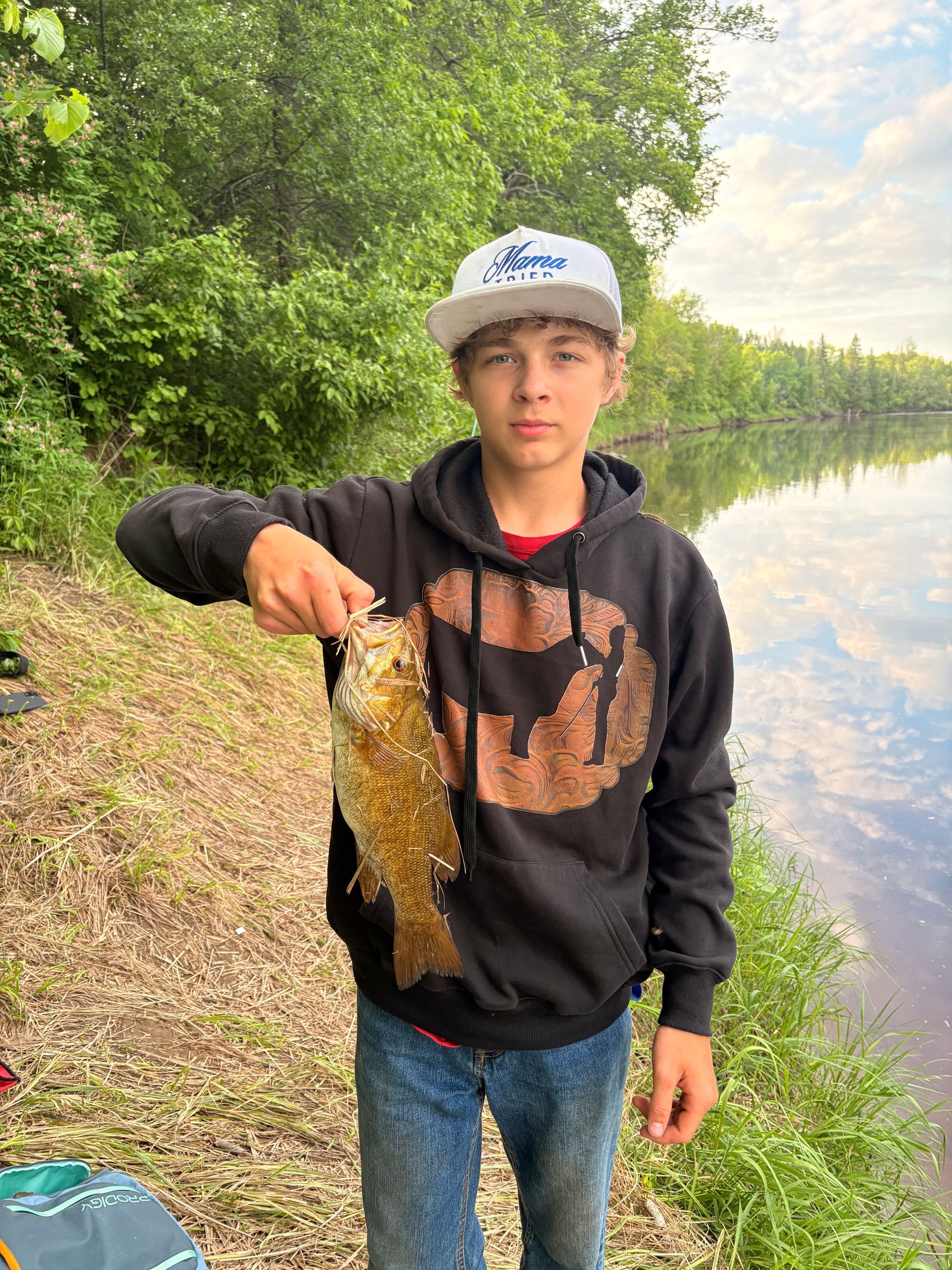 Person holding a fish by a riverbank, wearing a black hoodie and cap, with trees and water in the background.