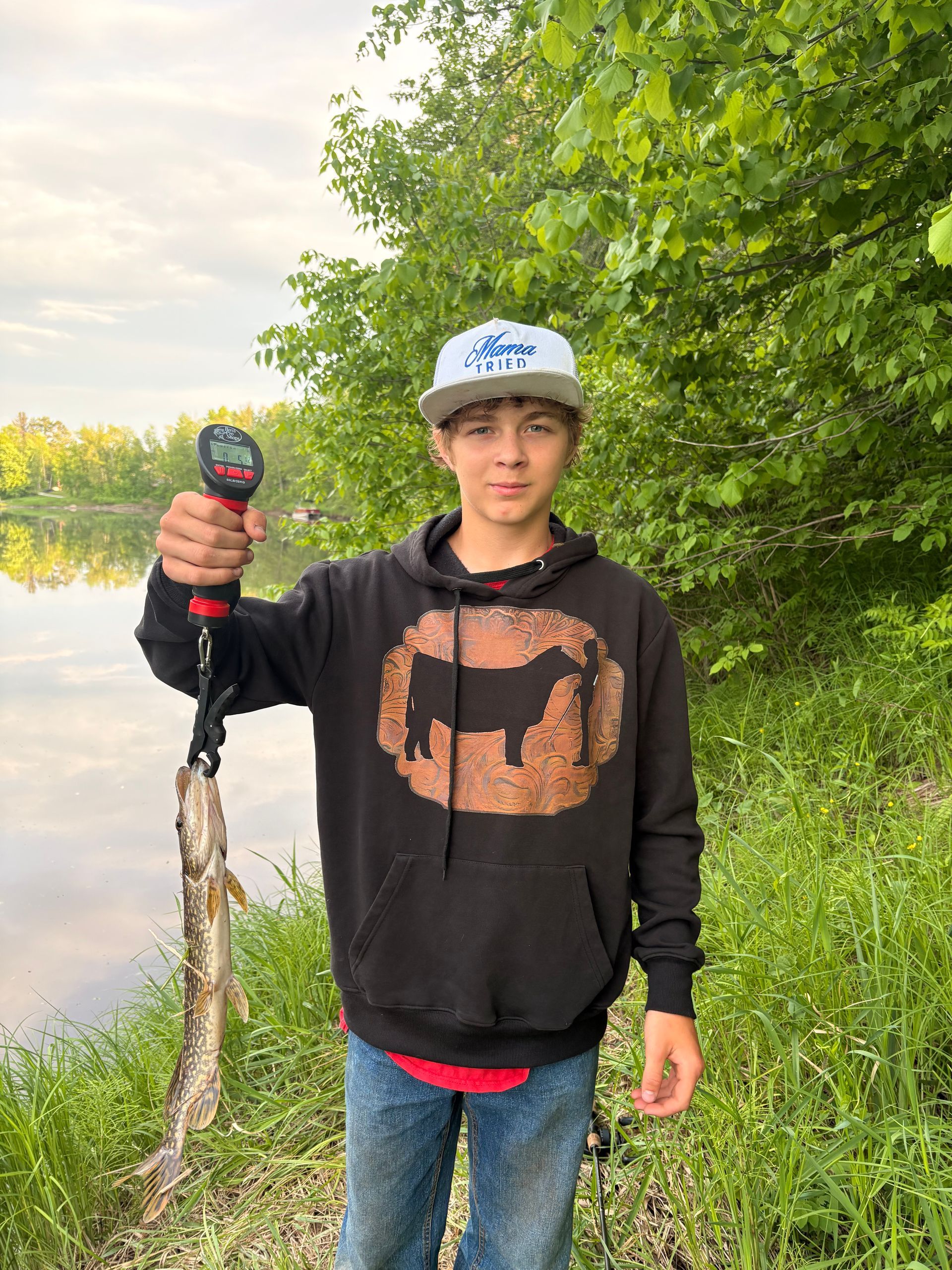 Person holding a fish by a lakeside, wearing a black hoodie and cap.