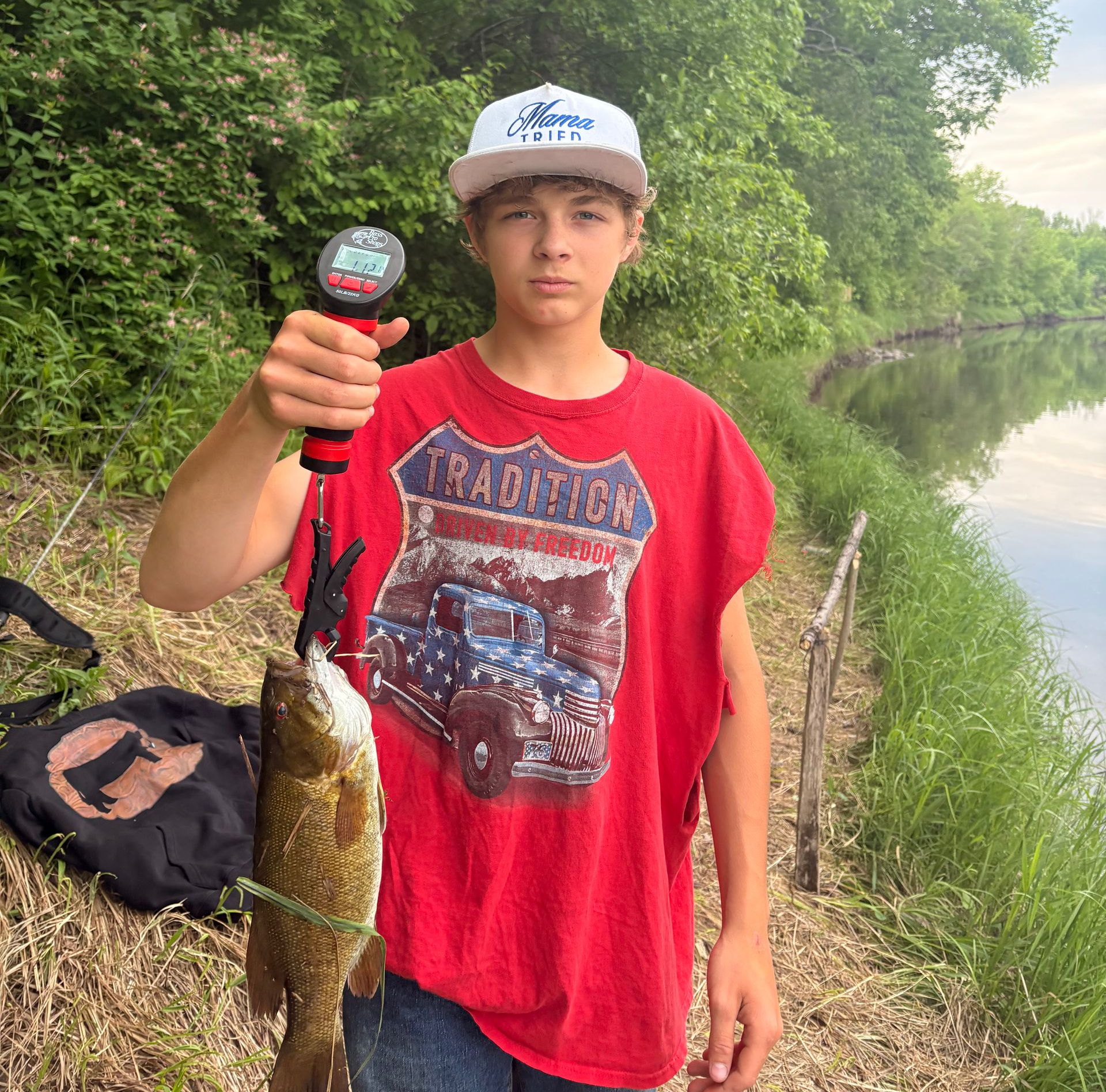 Young person in red shirt holding a fish by a riverbank, with trees and water in the background.