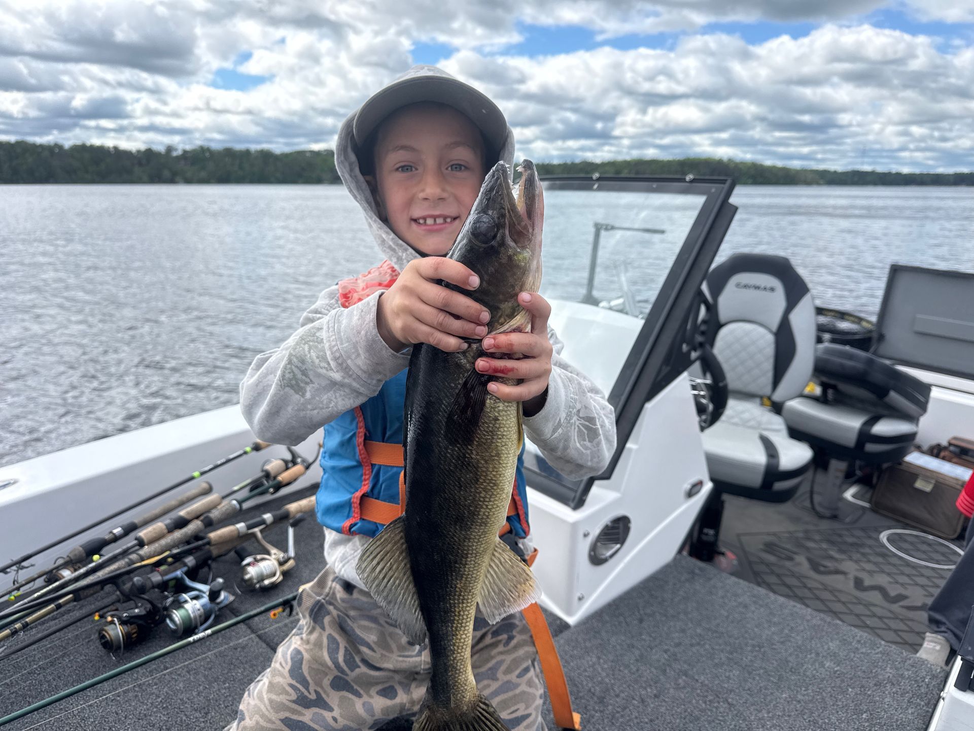 Child on a boat holding a large fish, with fishing rods and a lake in the background.