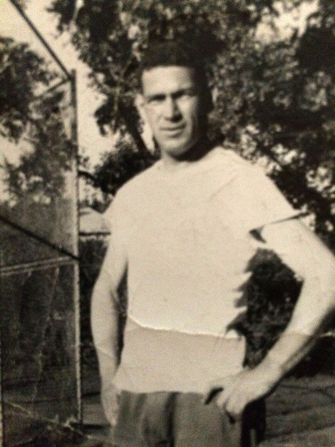 A man in a white shirt and dark pants stands outdoors with one hand on his hip.