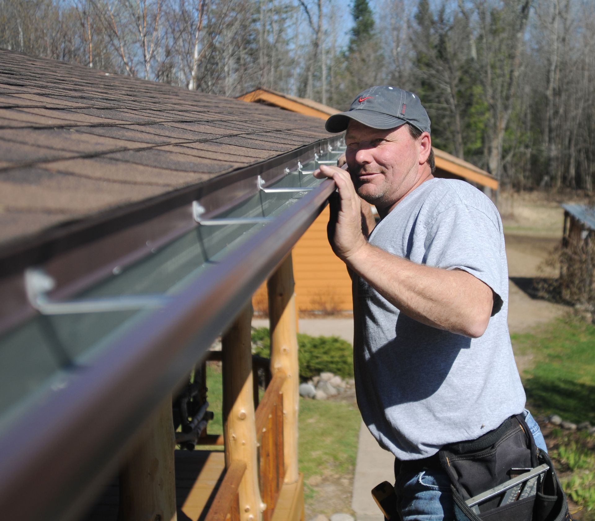 Worker installing gutter on a roof, leaning over the eaves with tools at his side.