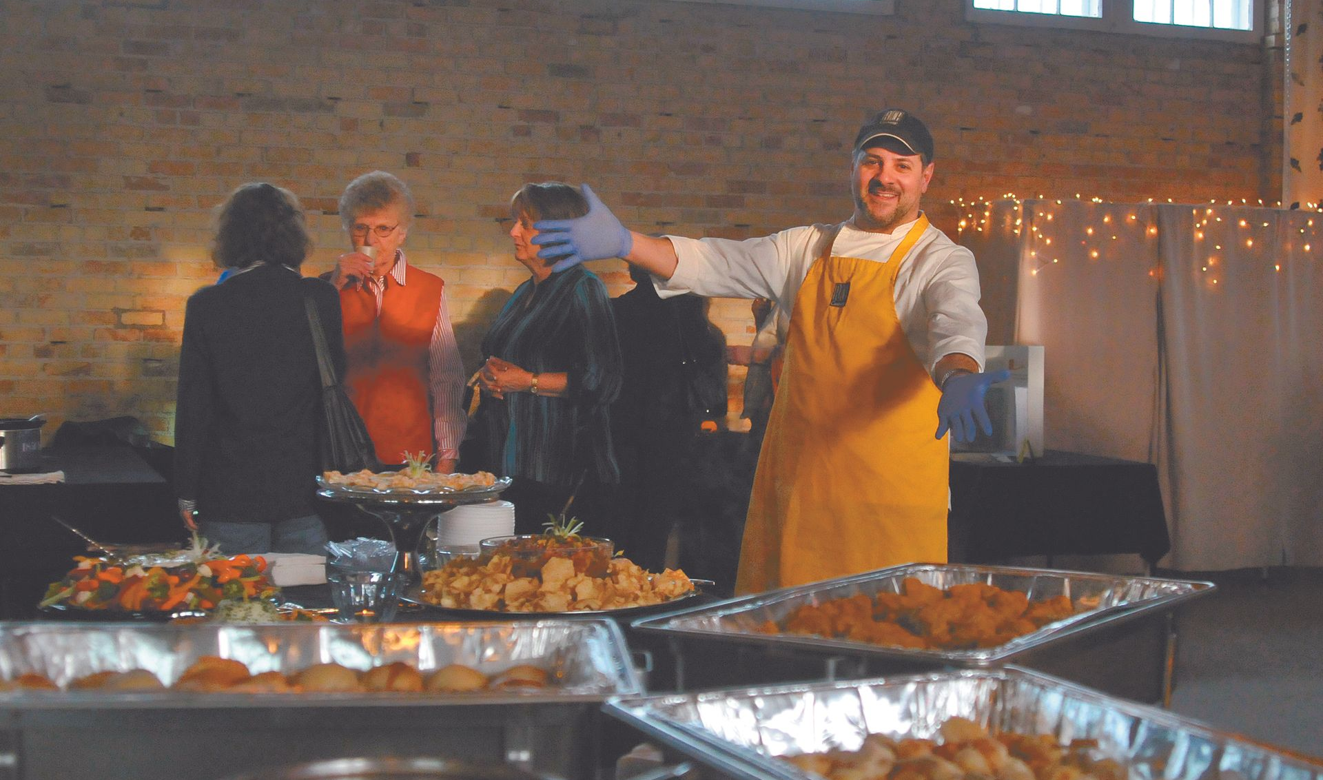 Buffet table with covered dishes and a man in a yellow apron serving guests in a rustic hall