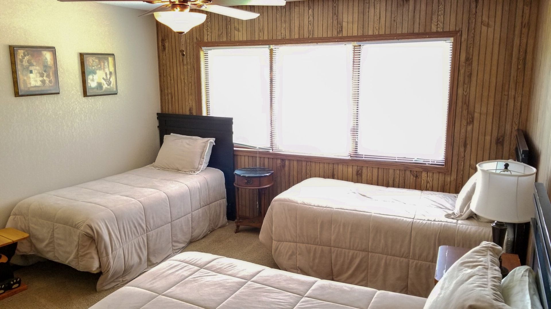 Two twin beds in a wood-paneled bedroom with a large window and ceiling fan.