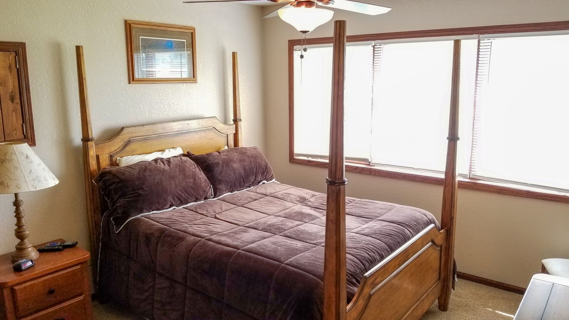 Bedroom with a four-poster bed, dark quilt, wooden nightstand, and bright window.