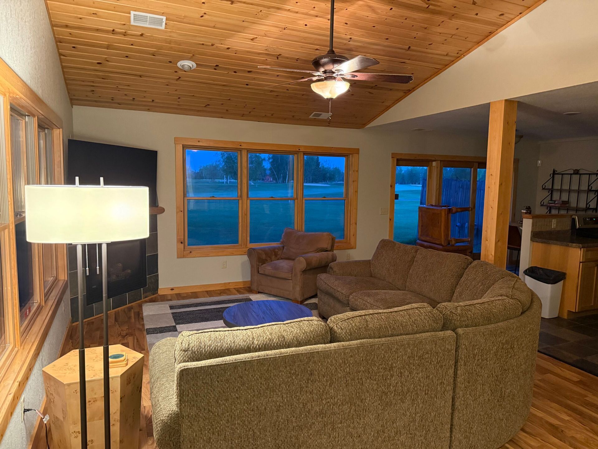 Cozy living room with brown sectional sofa, blue rug, floor lamp, large windows, and wood-paneled ceiling