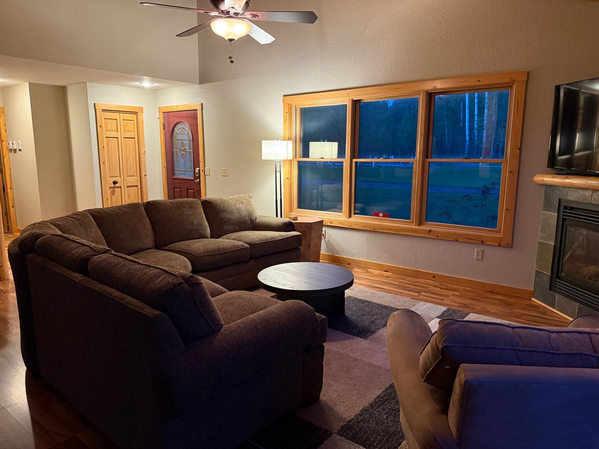 Cozy living room with brown sectional, coffee table, fireplace, lamp, and large window at dusk
