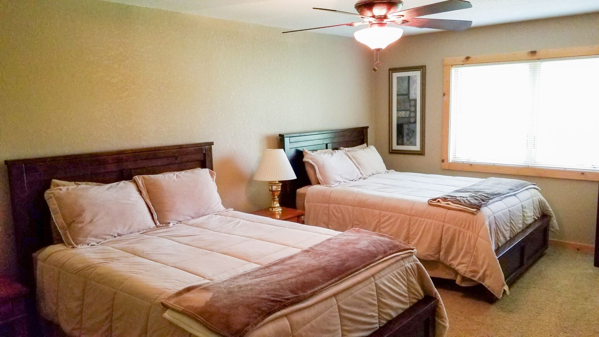 Bedroom with two beds, a ceiling fan, lamp, and large window letting in daylight